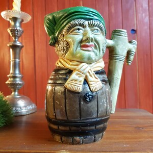 May include: A ceramic beer stein with a face, green hat, and scarf. The stein is barrel-shaped with a handle and a dark brown finish. The face is painted with green, red, and beige tones.