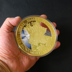 Puede incluir: Una moneda de color dorado que presenta un mapa de Francia con las palabras "Les Monuments Historiques" y "Liberté, Egalité, Fraternité" alrededor del borde.