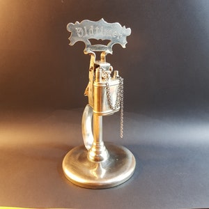 May include: Silver table lighter with a chain attached to the lighter. The lighter is on a silver stand with a sign that reads "Oldtimer".