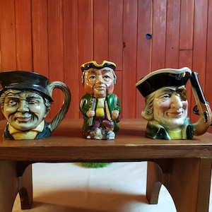 Puede incluir: Tres jarras de cerámica antiguas con forma de personajes sobre una mesa de madera. Las jarras representan a hombres con caras sonrientes y sombreros. Una jarra tiene un asa con forma de cara, y otra sostiene una pistola.