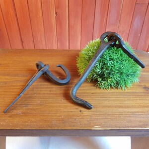 May include: Two antique, dark metal hooks on a wooden surface. One hook is a long, curved piece with a loop at the top. The other is a straight piece with a curved end. The hooks are displayed on a wooden table with a green, mossy accent.
