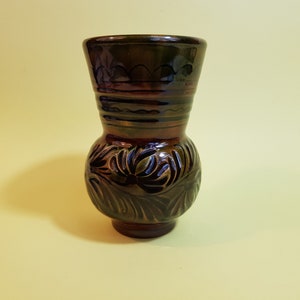 May include: A dark brown ceramic vase with a bulbous base and a narrower neck. The vase features carved floral patterns on the base and horizontal bands of color around the neck. The vase is approximately 7 inches tall.