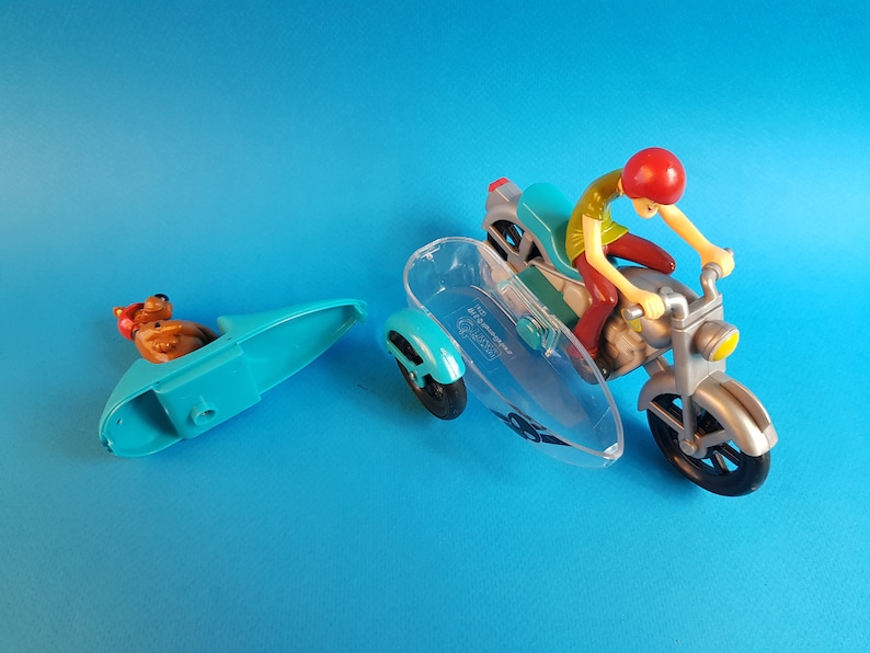 May include: A blue and silver toy motorcycle with a clear sidecar. A cartoon character with a red hat is riding the motorcycle. A separate blue sidecar with a cartoon dog figure is included.