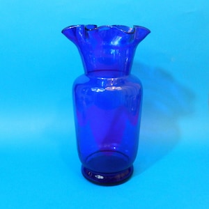 以下が含まれることがあります： コバルトブルーのガラスの花瓶。縁は波打っており、丸みを帯びた底と円筒形の胴体で、上部に向かって広がっています。透明なガラスを通して光が差し込みます。明るい青色の背景に置かれています。