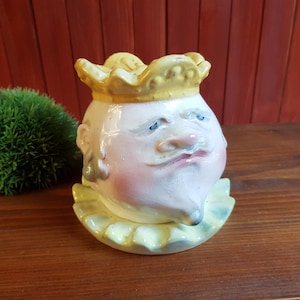 May include: A ceramic figurine of a king's head with a yellow crown and ruffled collar. The face is painted with pink cheeks, blue eyes, and a long nose. The figurine is on a wooden surface.