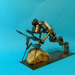 May include: A metal sculpture of a person doing a yoga pose, made from recycled materials like nuts, bolts, and spark plugs. The sculpture is on a metal base and is sitting on a rock.
