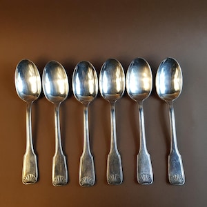 May include: Six silver spoons with ornate handles. The spoons are arranged in a row on a brown surface.