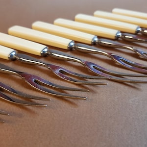 May include: A set of antique serving forks with ivory-colored handles and stainless steel tines. The forks are arranged diagonally, showcasing their elegant design and classic appeal. The handles are approximately 4 inches long.