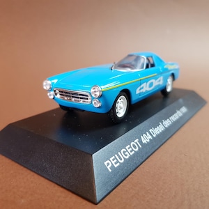 May include: A diecast model of a blue Peugeot 404 Diesel des records car. The model has the number "404" in white on the side and sits on a black display stand. The stand has the text "PEUGEOT 404 Diesel des records".
