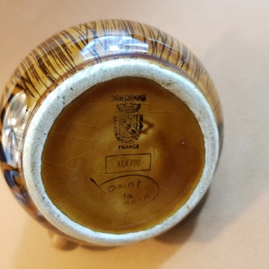 May include: A brown and tan ceramic pitcher with a striped pattern. The bottom of the pitcher has the text "SARREGUEMINES FRANCE" and "ACAJOU". The pitcher has a white rim and a small foot.