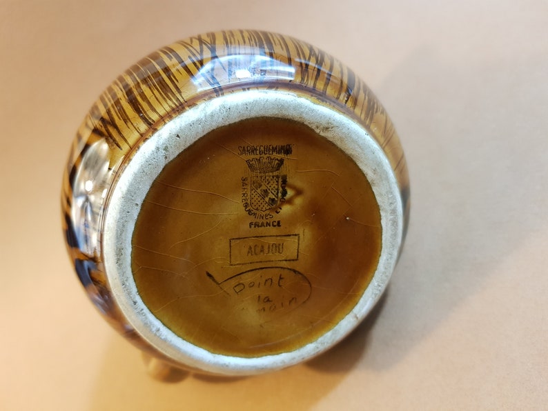 May include: A brown and tan ceramic pitcher with a striped pattern. The bottom of the pitcher has the text "SARREGUEMINES FRANCE" and "ACAJOU". The pitcher has a white rim and a small foot.