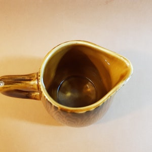 May include: A brown ceramic creamer with a handle and a spout. The creamer has a textured pattern on the exterior and a light brown interior. The handle has a decorative element on the end. The creamer is empty.