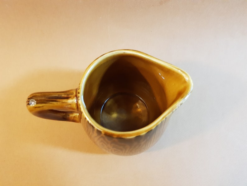 May include: A brown ceramic creamer with a handle and a spout. The creamer has a textured pattern on the exterior and a light brown interior. The handle has a decorative element on the end. The creamer is empty.