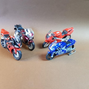 May include: Four toy motorcycles in red, blue, and maroon. The motorcycles have detailed features, including headlights, windshields, and racing graphics. The bikes are arranged on a neutral brown surface.