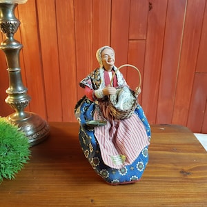 May include: A vintage figurine of a woman in traditional attire, holding a basket. The figurine wears a red and patterned shawl, a blue floral skirt, and a striped apron. The basket contains a white cloth. The figurine is on a wooden surface.