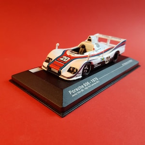 May include: A white, blue, and red Porsche 936 race car model with the number 20 on the side. The model is on a black display base with the text "Porsche 936 - 1976" and "Jacky Ickx (B) - Gijs Van Lennep (NL)".