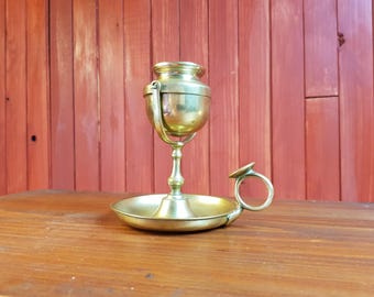 Puede incluir: Una lámpara de aceite de latón vintage. Presenta una base redonda, un tallo central y una parte superior en forma de copa. La lámpara tiene un asa y un pequeño anillo en el lateral. El fondo es una superficie de madera y una pared de madera roja.