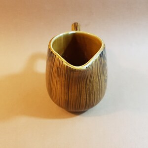 May include: A brown and tan ceramic creamer with a wood grain pattern. The creamer has a small handle and a spout for pouring. The interior is a lighter tan color. The creamer is approximately 5 inches tall.