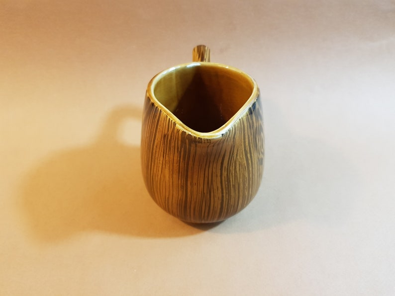 May include: A brown and tan ceramic creamer with a wood grain pattern. The creamer has a small handle and a spout for pouring. The interior is a lighter tan color. The creamer is approximately 5 inches tall.