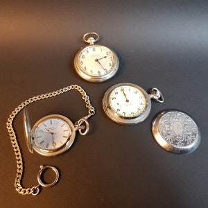 May include: Three silver pocket watches with white faces and black hands. One watch has a chain attached to it.