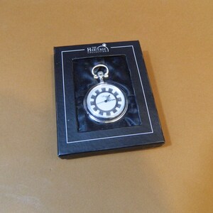 May include: A silver pocket watch with a black and white face, in a black box with the text "The Heritage Collection".