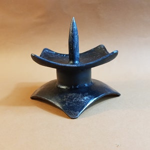 May include: A black metal candle holder with a pointed spike on top. The holder has a square base with rounded edges.