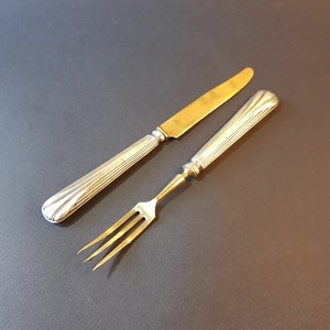 May include: A vintage knife and fork set. The knife has a gold blade and a silver handle with a ribbed design. The fork has a gold tines and a silver handle with a ribbed design. The set is likely made of silver or a silver alloy.