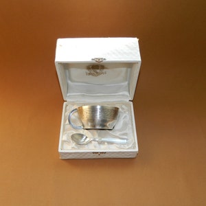 May include: A silver tea cup and spoon set presented in a white, quilted box. The cup has a handle and a perforated band around the top. The spoon is silver and rests beside the cup on a satin-like fabric.