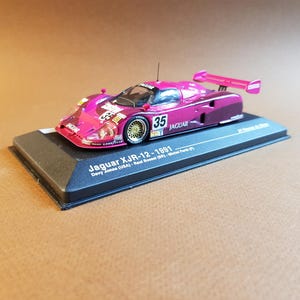 May include: A miniature magenta Jaguar XJR-12 race car model from 1991, with the number 35 on the side. The model sits on a black base with text that reads "Jaguar XJR-12-1991". The car has gold wheels and a rear wing.