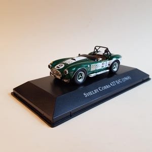May include: A green and white 1964 Shelby Cobra 427 S/C model car on a black display base. The car has a number "2" on the side and the text "SHELBY COBRA 427 S/C (1964)" on the base.
