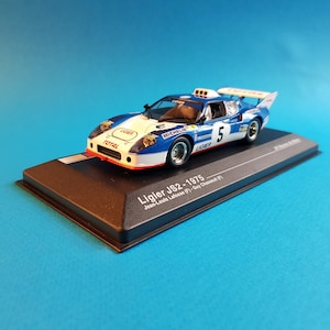 May include: A blue and white Ligier JS2 race car model with the number 5 on the side. The car is on a black display base with the text "Ligier JS2 - 1975 Jean-Louis Lafosse (F) - Guy Chasseuil (F)".