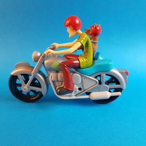 May include: A silver toy motorcycle with a red-helmeted figure riding and a brown figure sitting behind. The motorcycle has a blue seat and a white exhaust pipe.