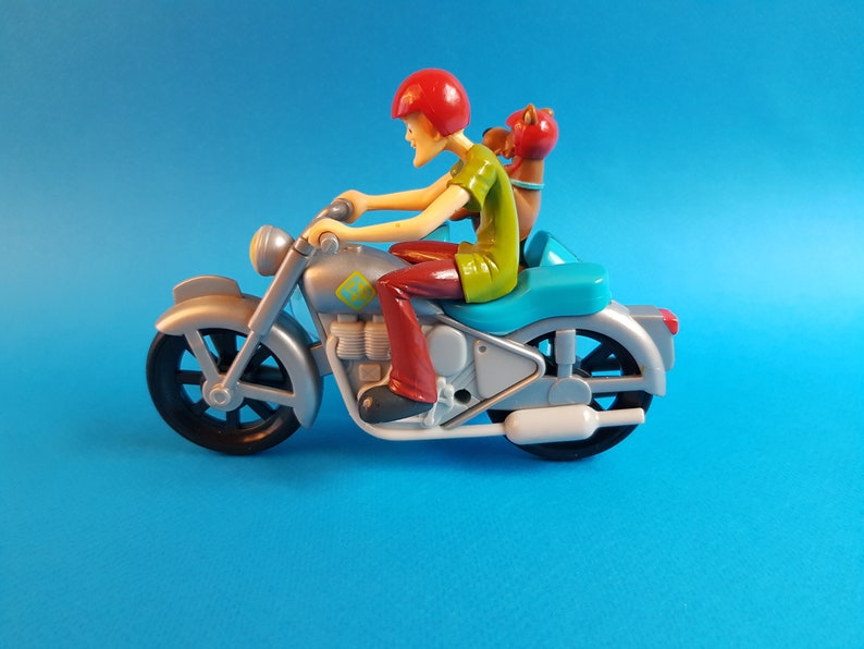 May include: A silver toy motorcycle with a red-helmeted figure riding and a brown figure sitting behind. The motorcycle has a blue seat and a white exhaust pipe.