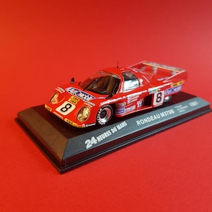 May include: A red, white, and blue 1:43 scale model car of a Rondeau M379B race car. The car is on a black display base with the text "24 Heures du Mans" and "RONDEAU M379B 1981" printed on it.
