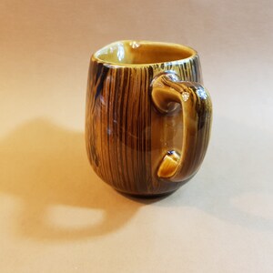 May include: A ceramic mug with a wood grain design. The mug is brown with vertical dark brown stripes and a curved handle. The interior of the mug is a lighter brown color. The mug is sitting on a light brown surface.