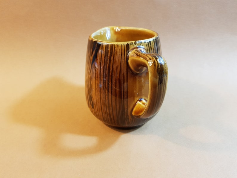 May include: A ceramic mug with a wood grain design. The mug is brown with vertical dark brown stripes and a curved handle. The interior of the mug is a lighter brown color. The mug is sitting on a light brown surface.