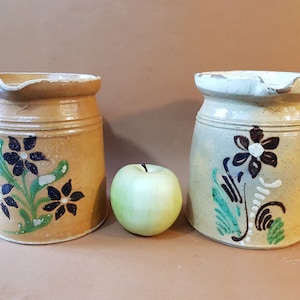 May include: Two vintage ceramic jars with floral designs. The jars are brown and white with black and green painted flowers. A green apple sits between the jars.
