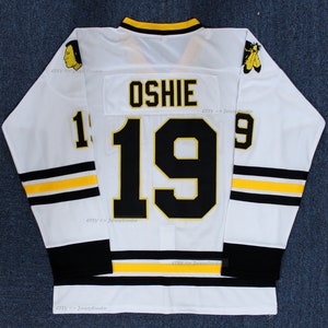 May include: A white hockey jersey featuring the name OSHIE and the number 19 in black with yellow trim on the back. The sleeves have black and yellow stripes, and the shoulders display circular logos. The jersey has a black and yellow collar.