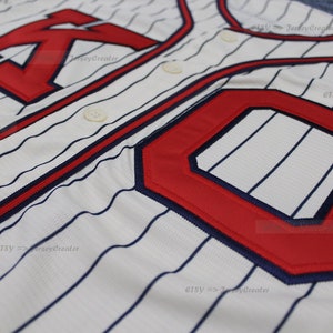 May include: White baseball jersey with blue and red stripes. The jersey has a red "K" and a red "C" on the front.