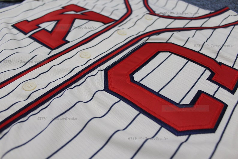 May include: White baseball jersey with blue and red stripes. The jersey has a red "K" and a red "C" on the front.