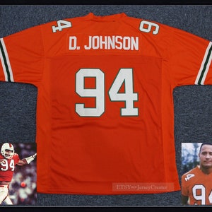 Può includere: Maglia da football arancione con numeri bianchi e bordini verdi. La maglia ha il nome "D. JOHNSON" e il numero "94" sul retro. Una foto di un giocatore di football che indossa la stessa maglia si trova nell'angolo in basso a destra.