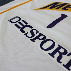 May include: White athletic jersey with a purple number "1" and the text "DECSPORT" in purple on a white patch. The jersey has a yellow stripe on the shoulder and the text "METS" in yellow on the chest.