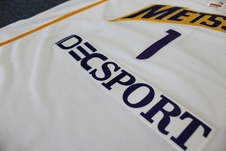 May include: White athletic jersey with a purple number "1" and the text "DECSPORT" in purple on a white patch. The jersey has a yellow stripe on the shoulder and the text "METS" in yellow on the chest.