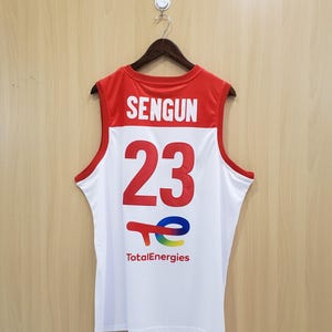 K&ouml;nnte beinhalten: Wei&szlig;es und rotes Basketballtrikot mit dem Namen "SENGUN" und der Nummer "23" in gro&szlig;en roten Buchstaben. Das TotalEnergies-Logo befindet sich unten. Das Trikot h&auml;ngt an einem Holzb&uuml;gel.
