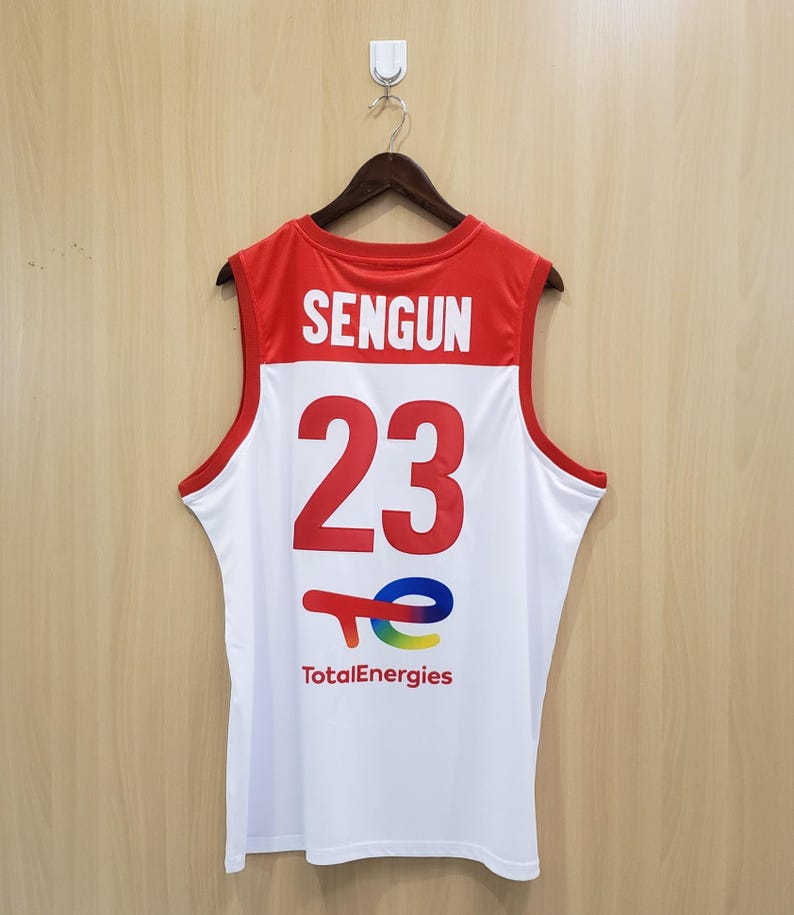 K&ouml;nnte beinhalten: Wei&szlig;es und rotes Basketballtrikot mit dem Namen "SENGUN" und der Nummer "23" in gro&szlig;en roten Buchstaben. Das TotalEnergies-Logo befindet sich unten. Das Trikot h&auml;ngt an einem Holzb&uuml;gel.