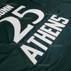 Puede incluir: Primer plano de una camiseta verde oscuro con letras y n&uacute;meros blancos. El n&uacute;mero "25" se muestra de forma destacada, con el nombre "DUNN" encima. Tambi&eacute;n se ve la palabra "ATHENS". La camiseta tiene una textura de malla.