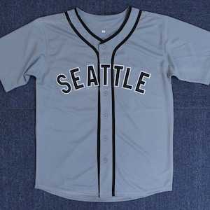 Puede incluir: Una camiseta de béisbol gris con ribetes negros y la palabra "SEATTLE" en letras negras en la parte delantera.