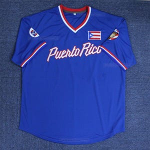 May include: A blue baseball jersey with the words "Puerto Rico" in white script across the chest. The jersey has a V-neck with red and white trim, and the Puerto Rican flag is on the chest. The sleeves have red and white stripes.