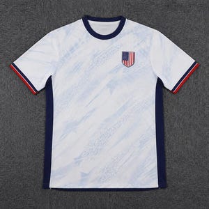 May include: A white short-sleeved t-shirt featuring a subtle light blue diagonal pattern across the front. The shirt has a dark blue crew neckline, dark blue side panels, and sleeve cuffs accented with thin red and dark blue stripes. A shield-shaped patch with the United States flag is positioned on the upper left chest.