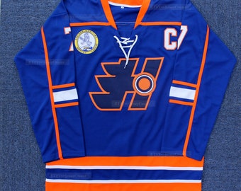 Puede incluir: Una camiseta de hockey azul con rayas naranjas y blancas. La camiseta tiene un gran logotipo naranja y negro en la parte delantera con el número "C7" en el hombro derecho.